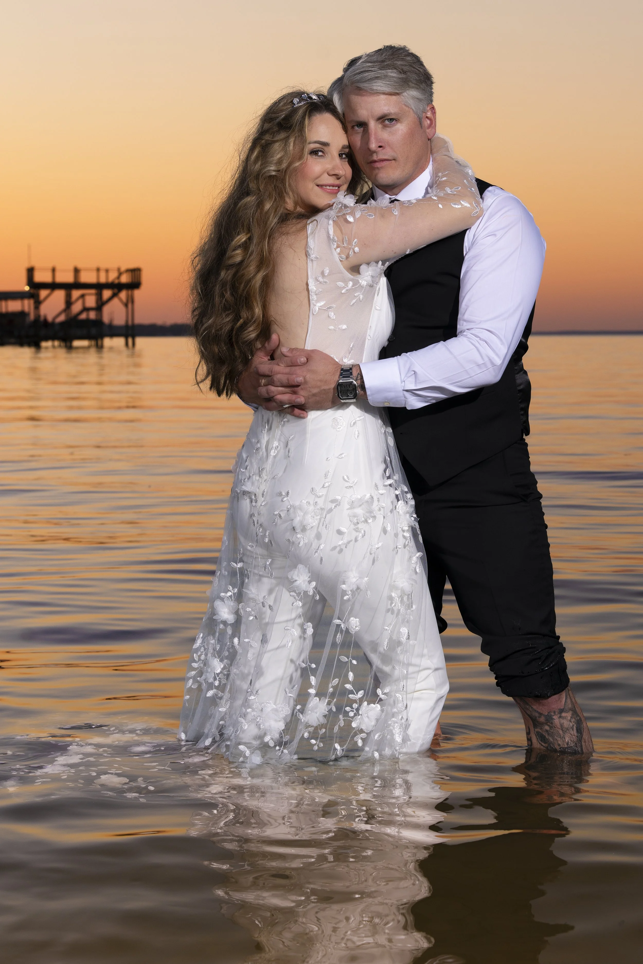 A couple in wedding attire, standing in water at sunset, embracing each other. The woman is in a white wedding dress with floral details, and the man in a white shirt with a black vest, with his hand on her waist. The water reaches their ankles, and a wooden dock extends out over the water in the background.