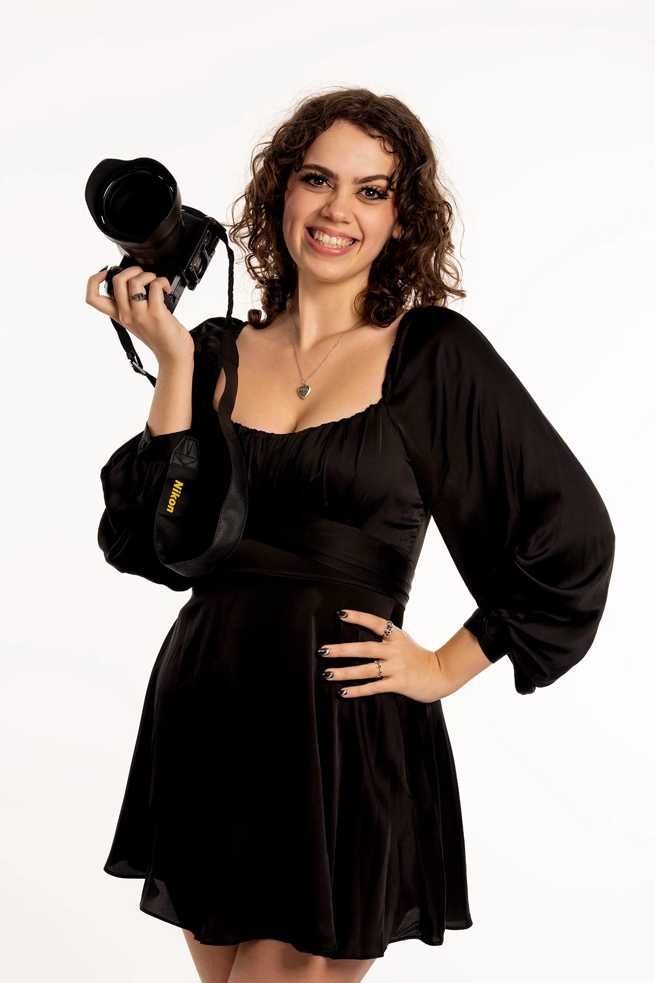 A young woman with curly brown hair, wearing a black dress with balloon sleeves, smiling and holding a Nikon camera in her right hand against a plain white background.