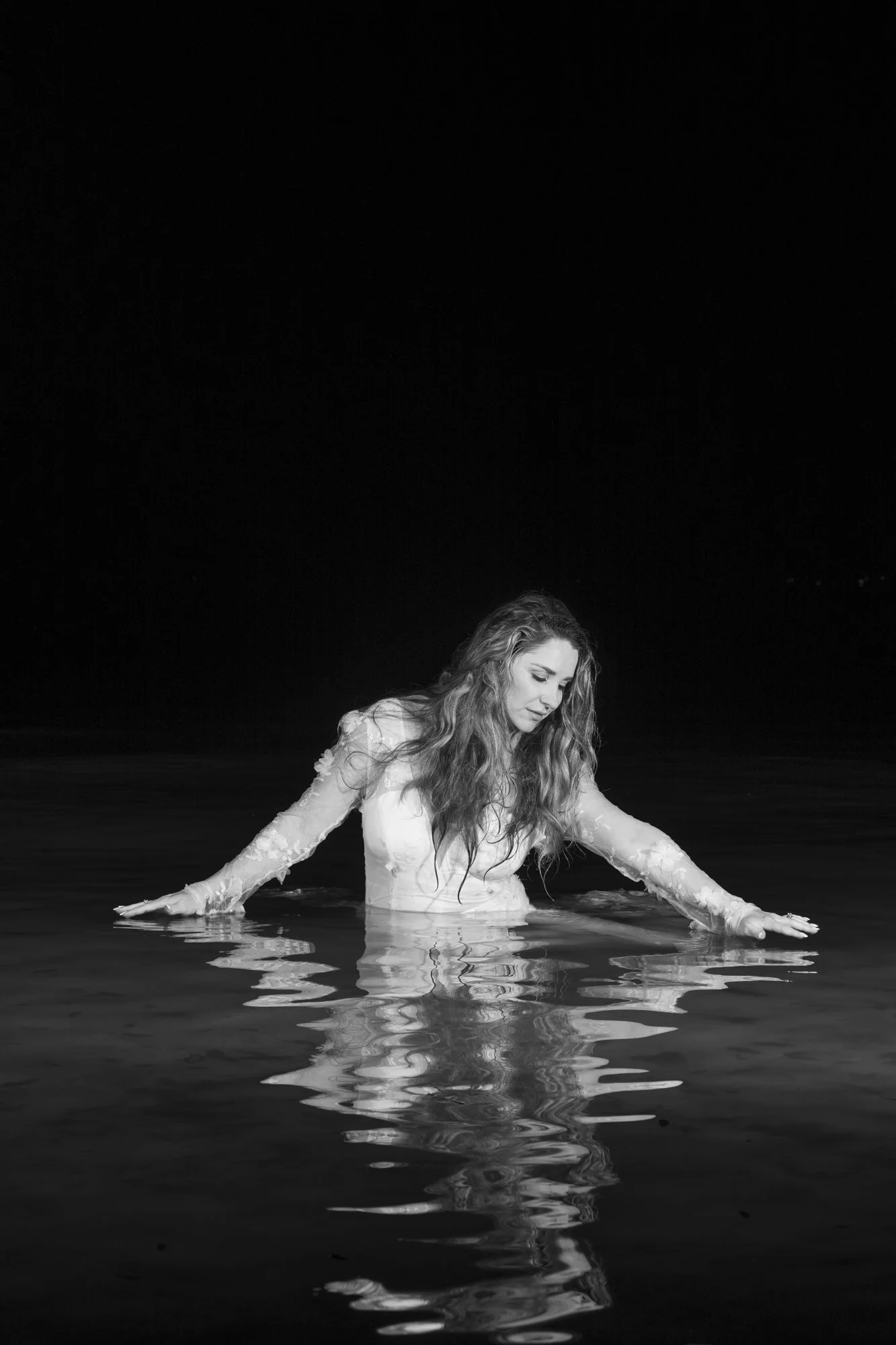 A woman with long wavy hair submerged in water up to her chest, wearing a lacy long-sleeve dress, with her arms stretched out to the sides and eyes closed in a serene pose against a dark background.