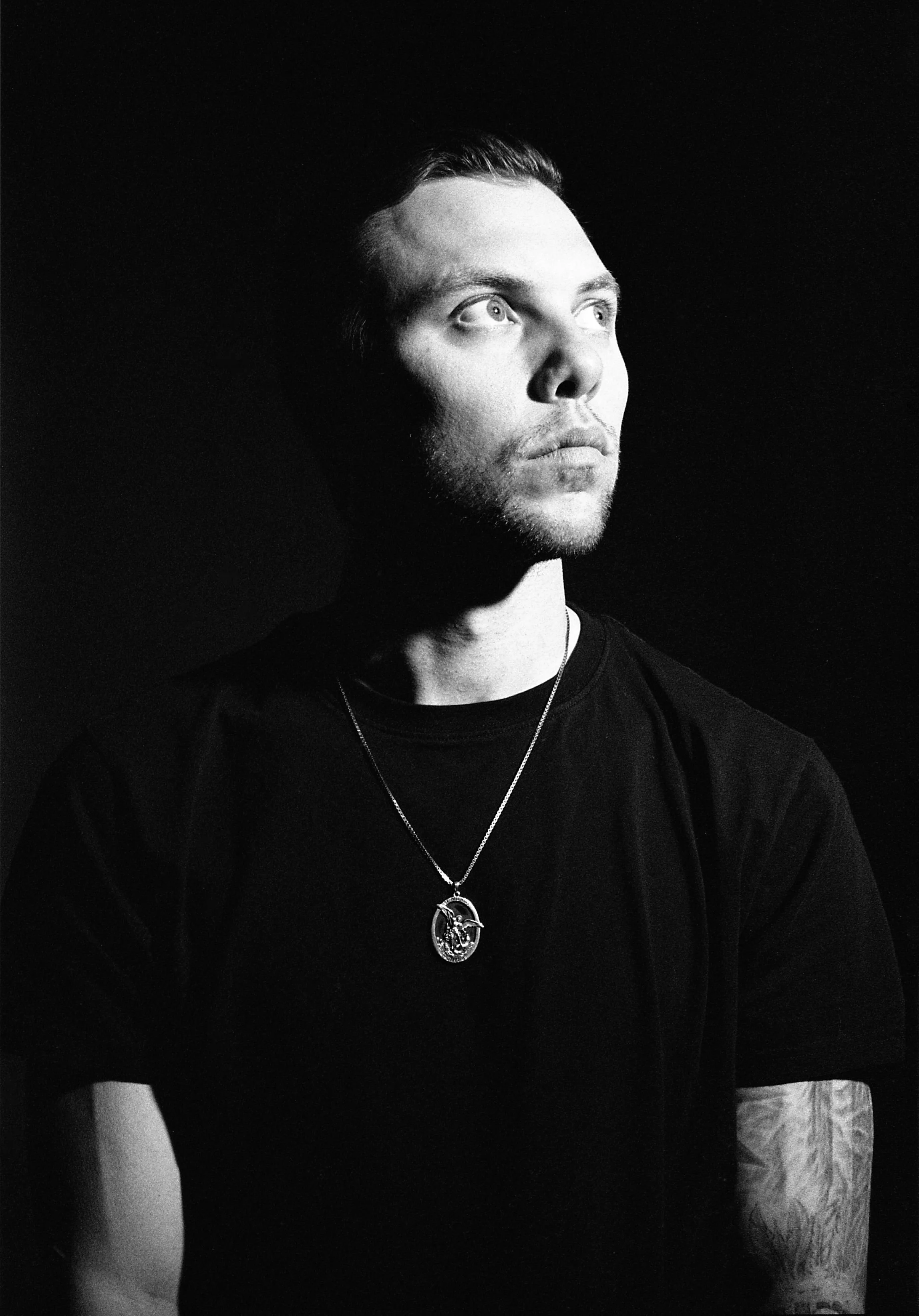 Black and white portrait of a young man with short hair, looking upward with a serious expression, wearing a chain with a pendant and a black shirt.