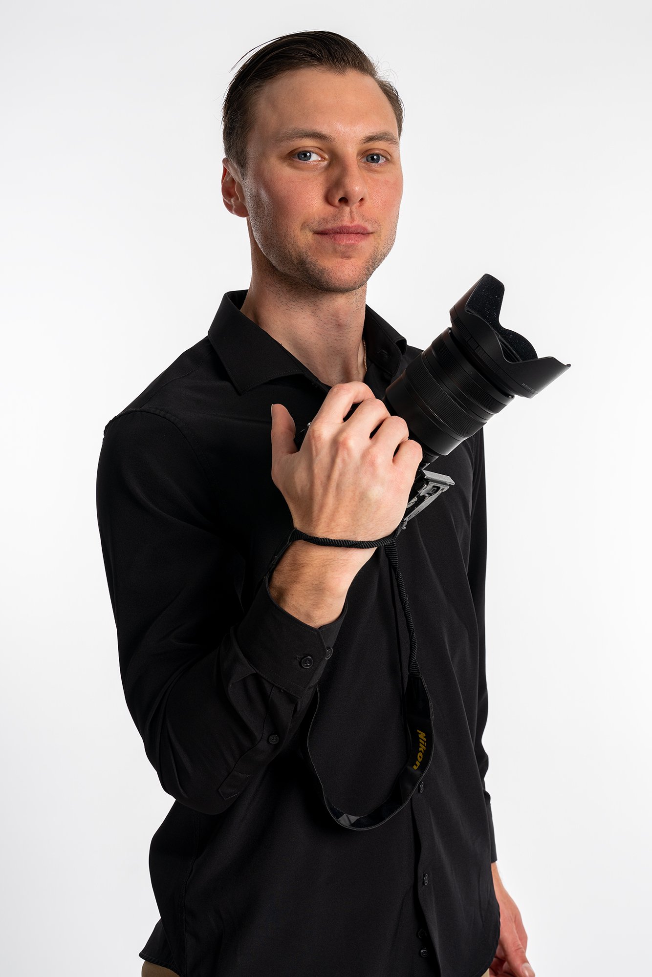 A young man with short brown hair, wearing a black shirt, holding a professional camera with a large lens, standing against a plain white background.