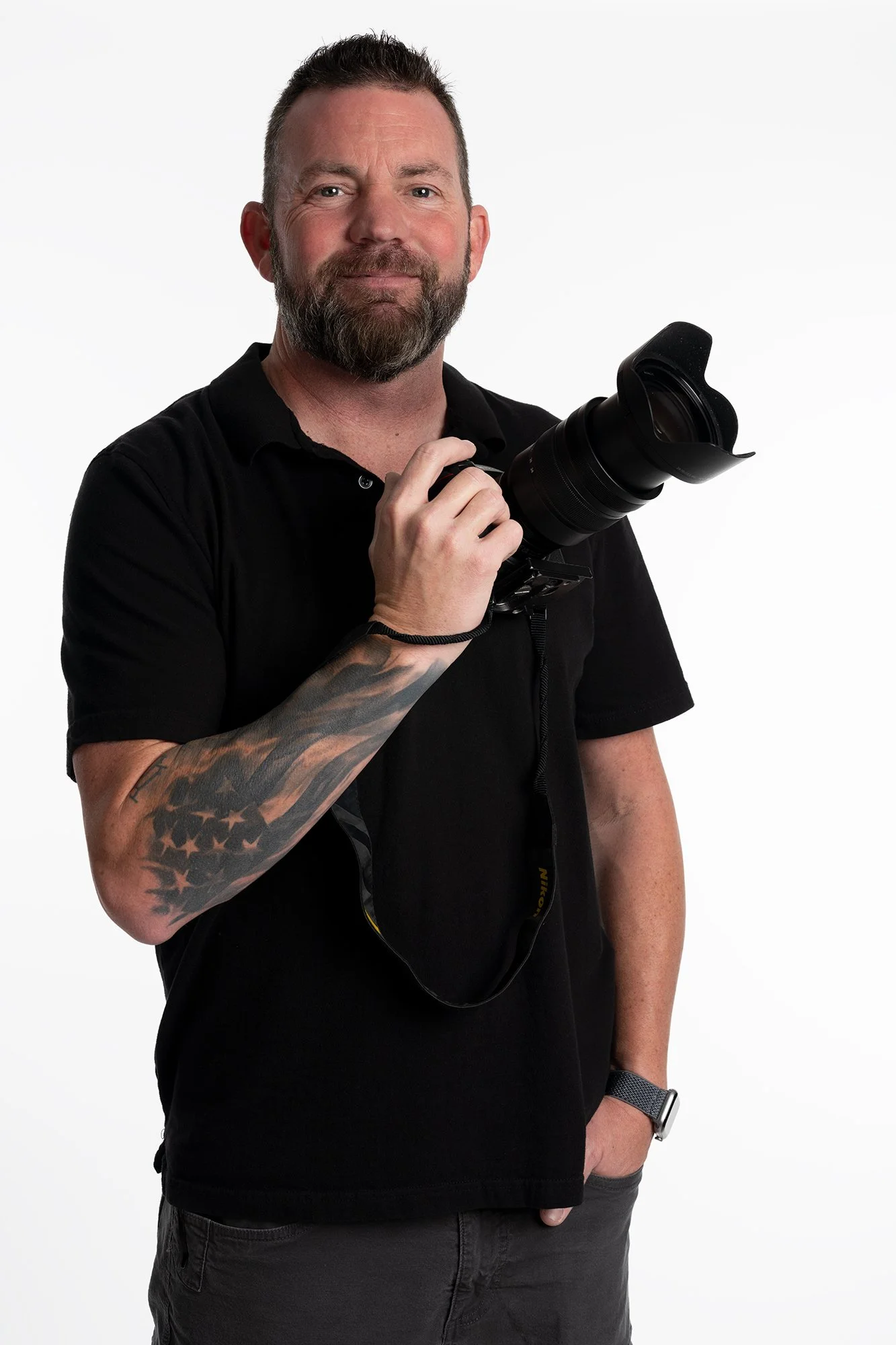 Man with a beard and tattooed arm holding a professional camera, wearing a black shirt and wristwatch, standing against a white background.