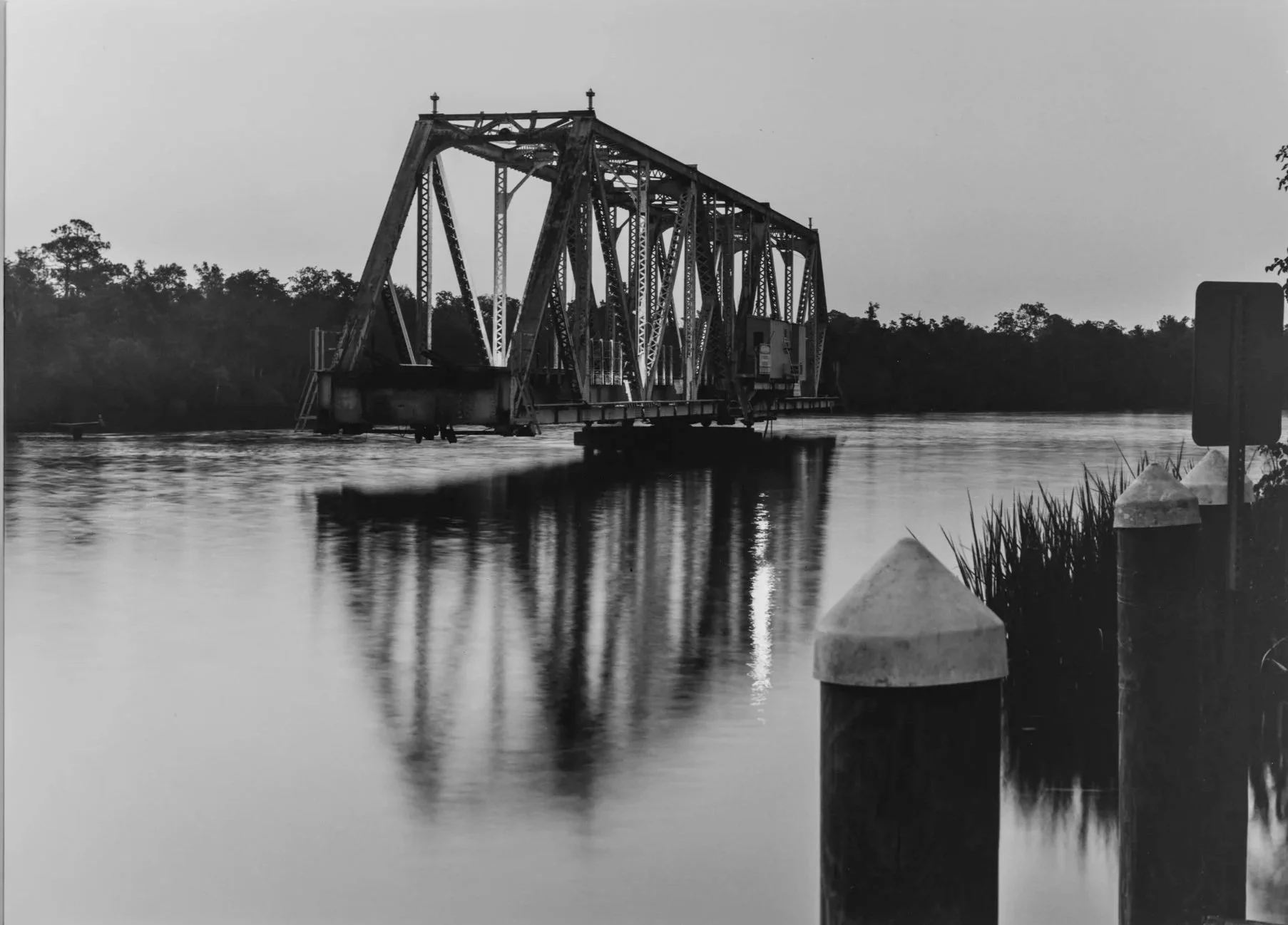 Black and white photo of an old bridge crossing a body of water, with trees in the background and posts along the water's edge in the foreground.