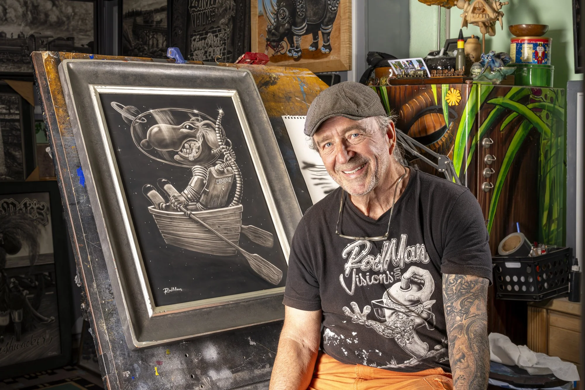 An older man with a gray beard and tattoos, smiling, sitting in an art studio. He wears a brown cap, black t-shirt, and orange pants. Behind him is a large black and white artwork of a mouse in a boat with sci-fi elements. The studio is decorated with colorful artworks, art supplies, and various decorative items.