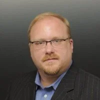 A man with blonde hair, glasses, and a beard, wearing a dark pinstripe suit jacket and a light blue shirt, against a dark gradient background.