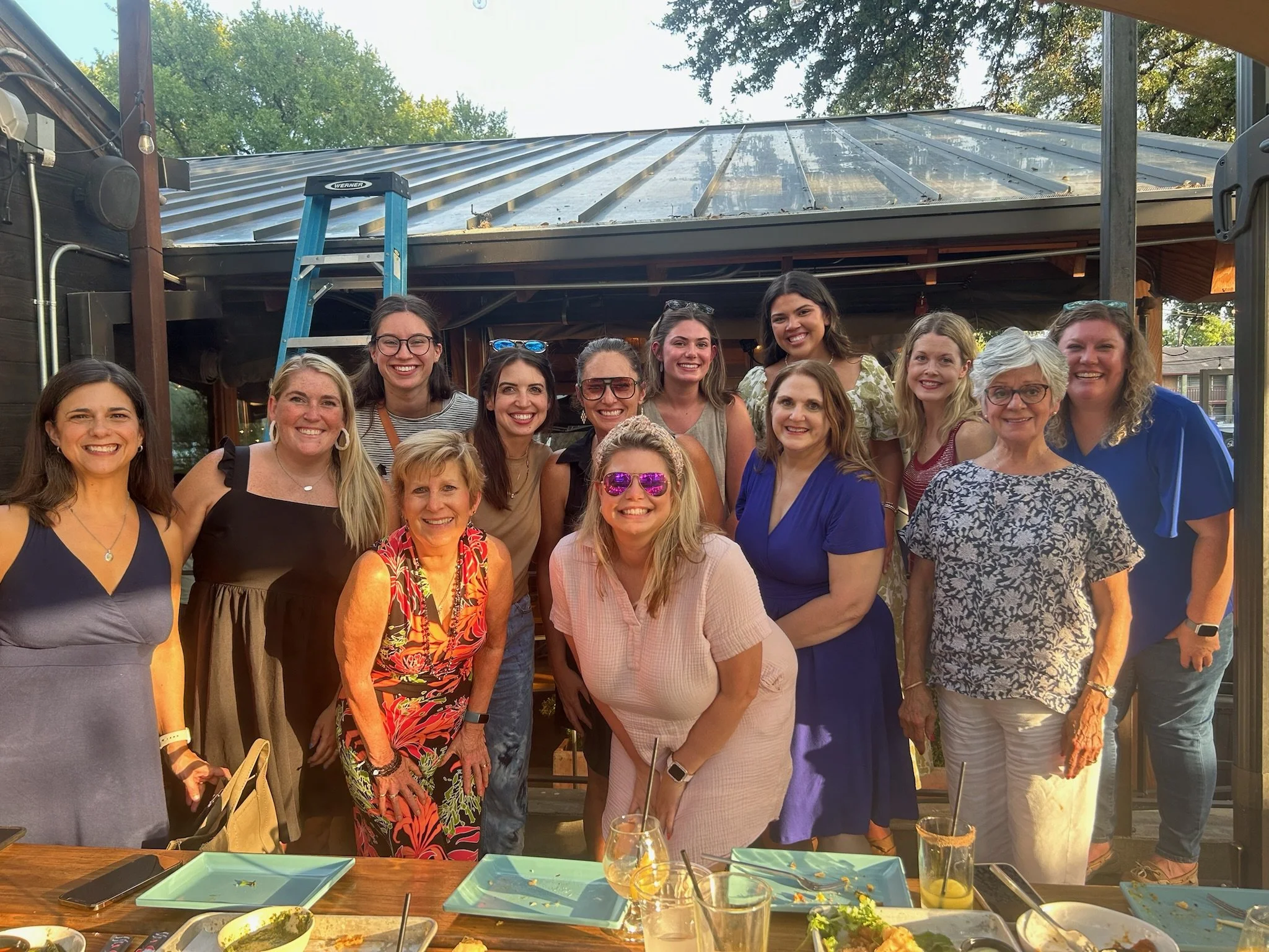 Group of women gathered outdoors on a patio, smiling for a photo under a metal and glass roof with green trees in the background. There are plates and drinks on the table in front of them.