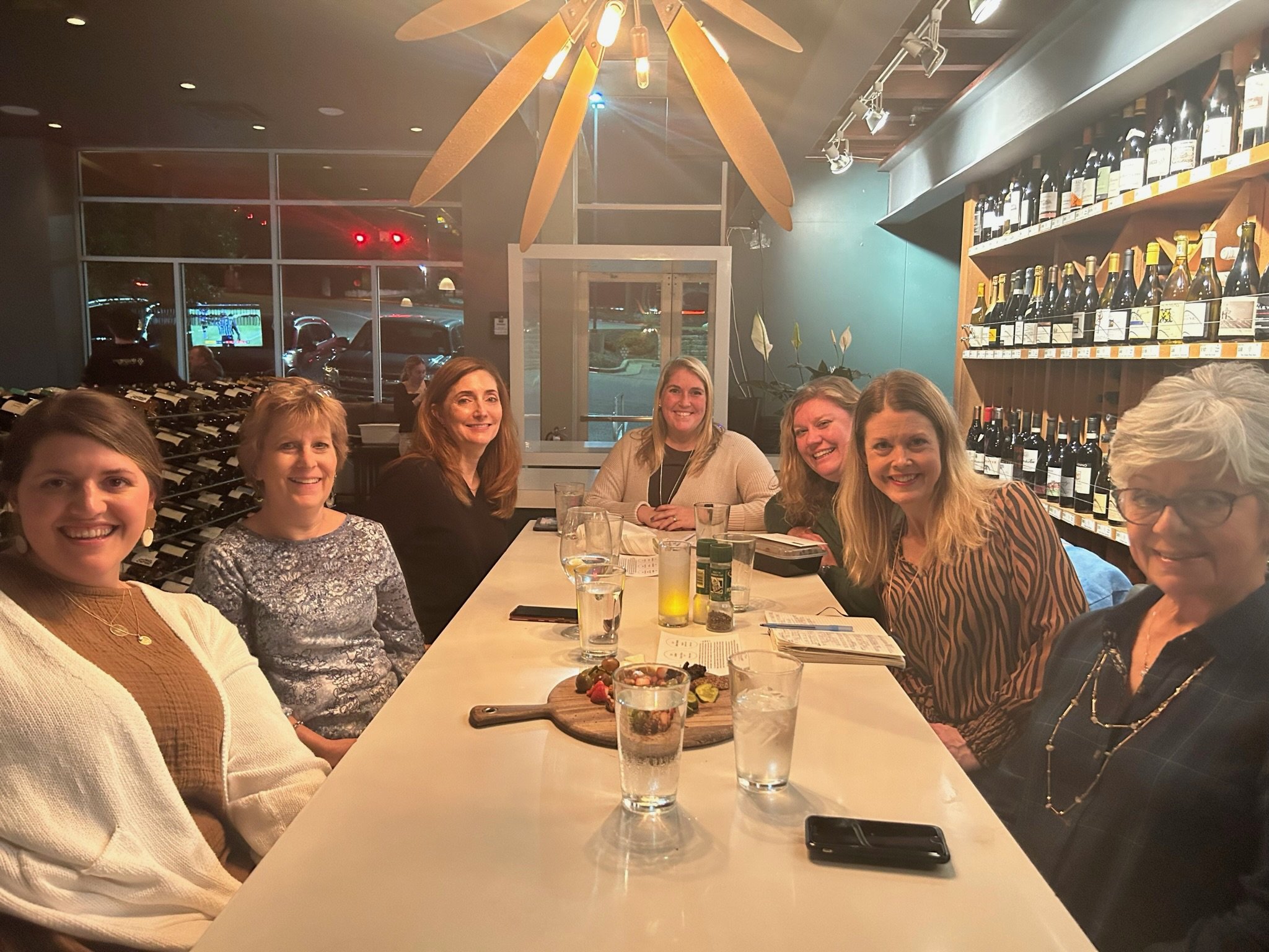 A group of seven women sitting around a long restaurant table, smiling for the camera, inside a cozy restaurant with wine bottles on shelves and a ceiling fan overhead.