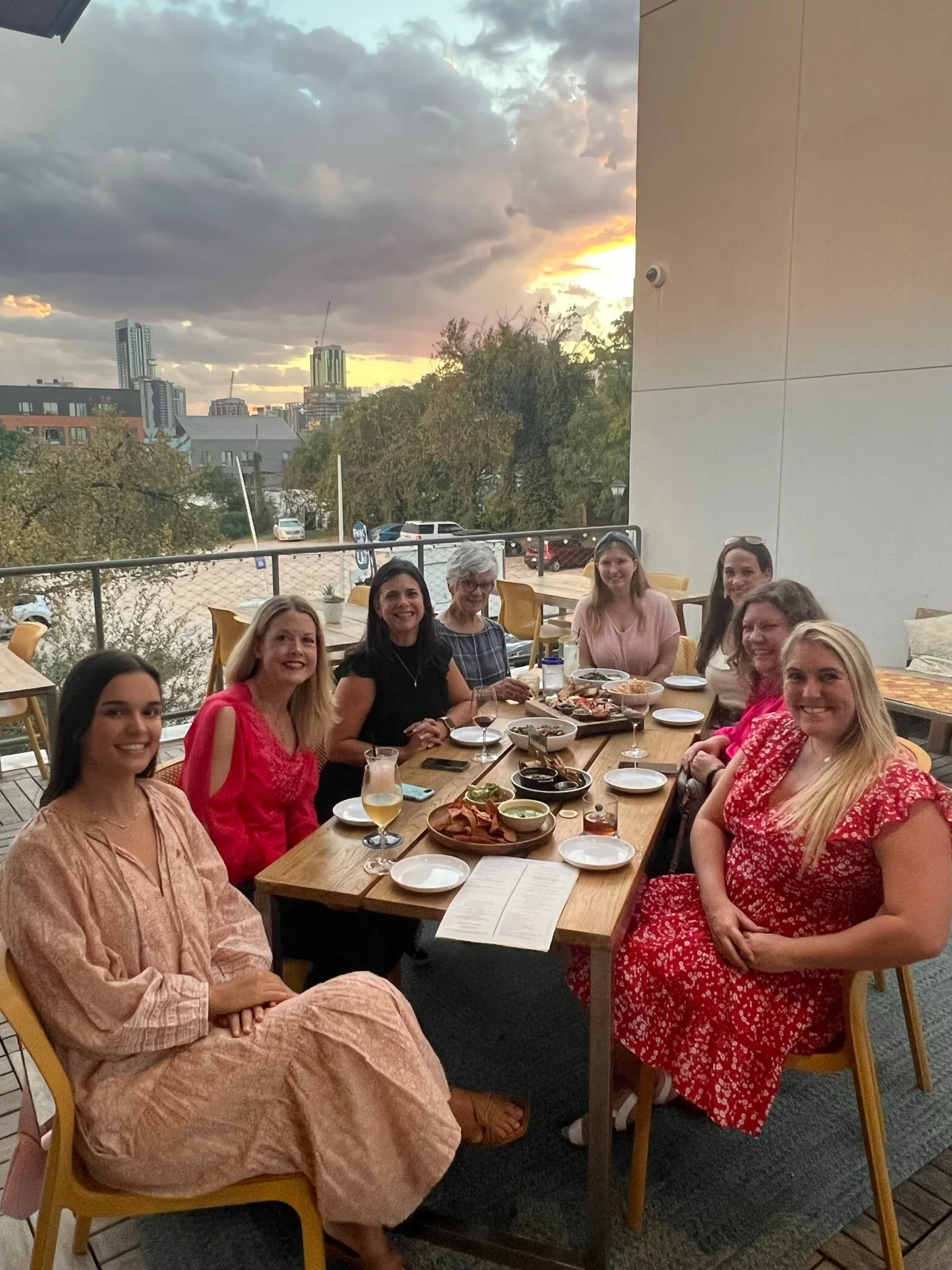 Group of women sitting around a table on a restaurant patio, enjoying food and drinks during sunset.