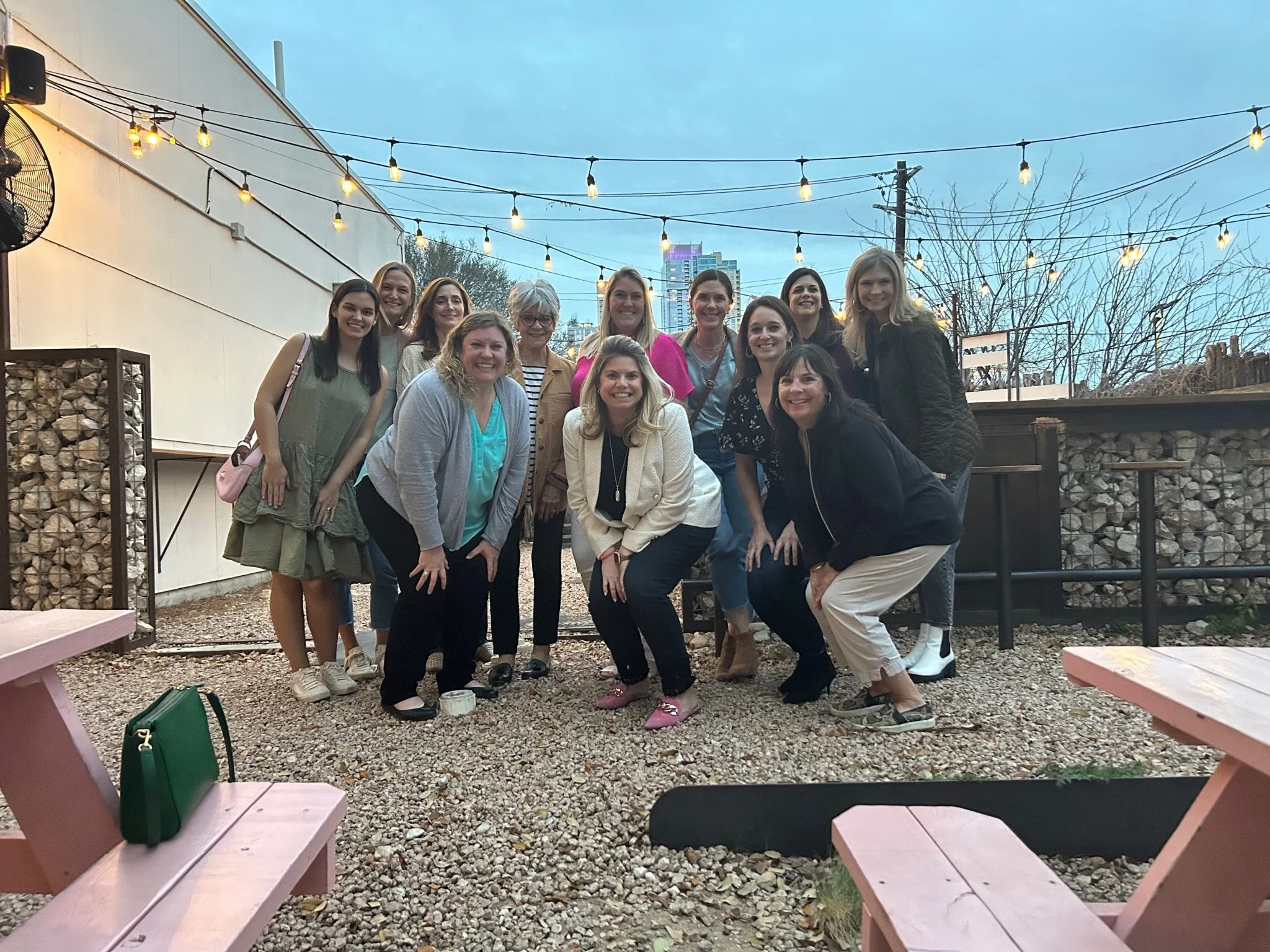 Group of women gathered outdoors on an evening with string lights overhead, standing and kneeling on a gravel surface. Pink picnic tables are visible in the foreground.
