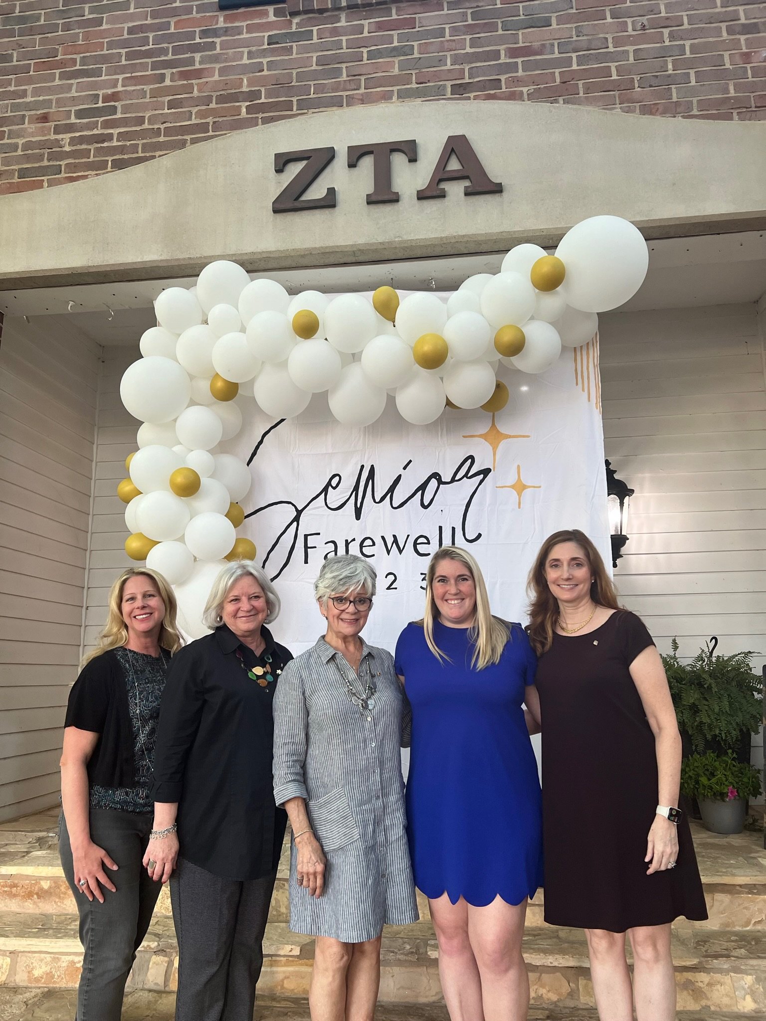 Five women standing on steps in front of a farewell event banner with white and gold balloons and the letters 'ZTA' on a building.