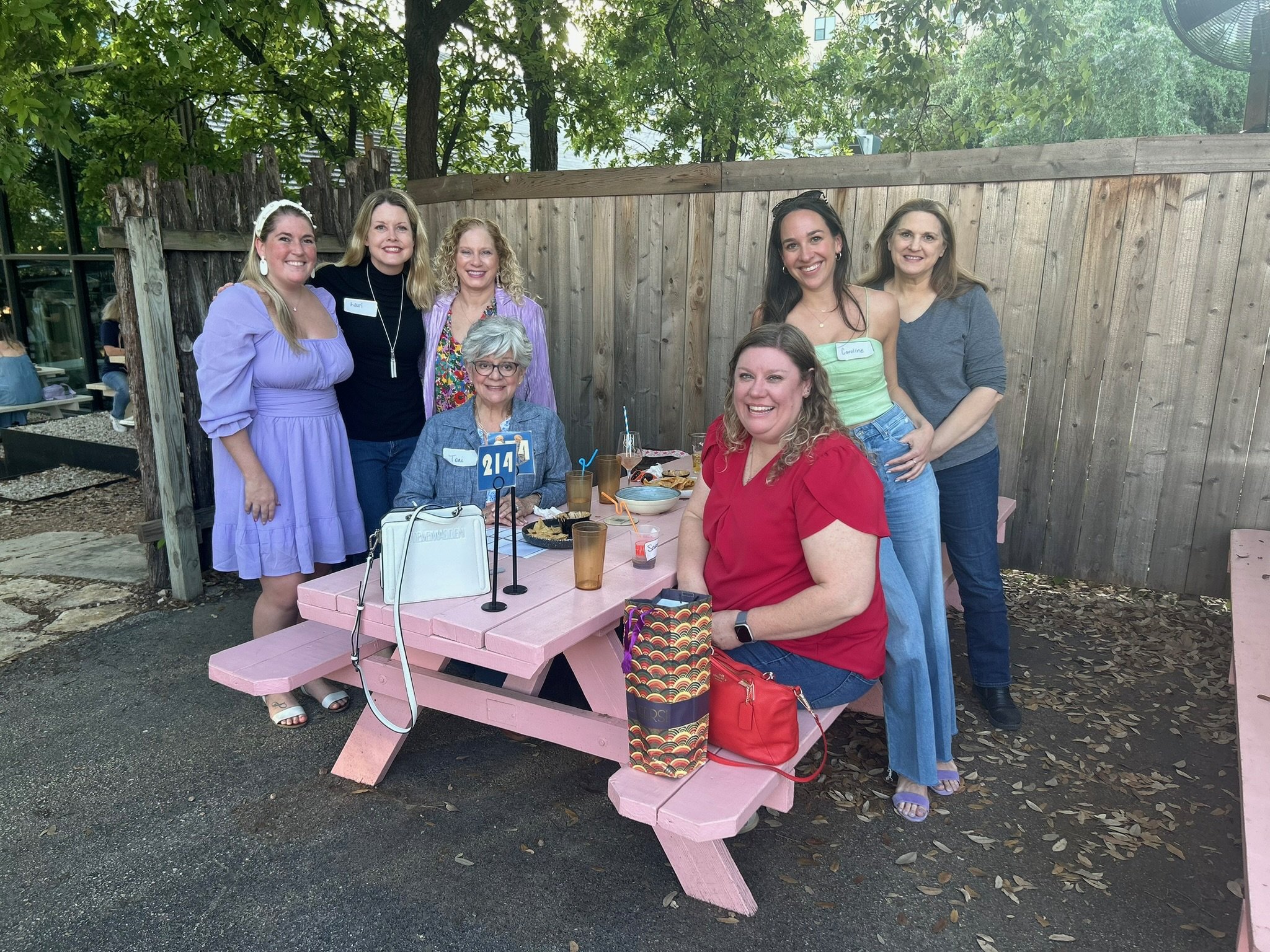 A group of eight women gathering around a pink picnic table outdoors, smiling for the photo, with some standing and one sitting at the table, which has food, drinks, and party decorations.