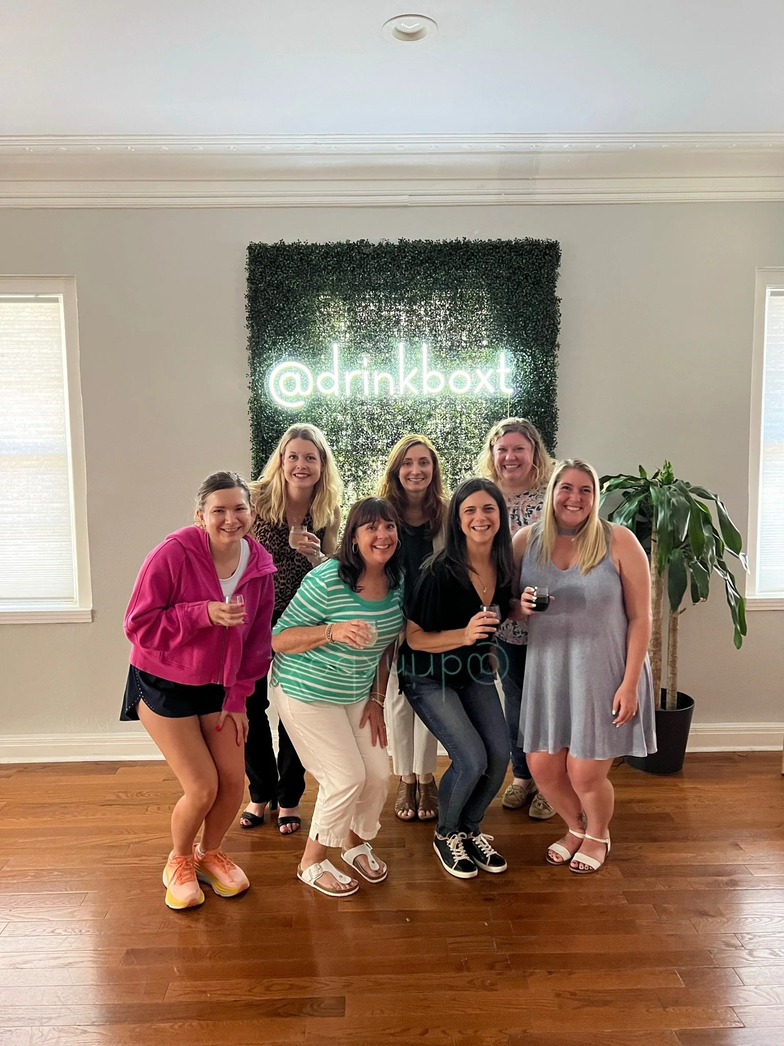 Group of eight women smiling and holding drinks in a room with a decorative green wall and neon text '@drinkboxt' behind them, next to a large potted plant.