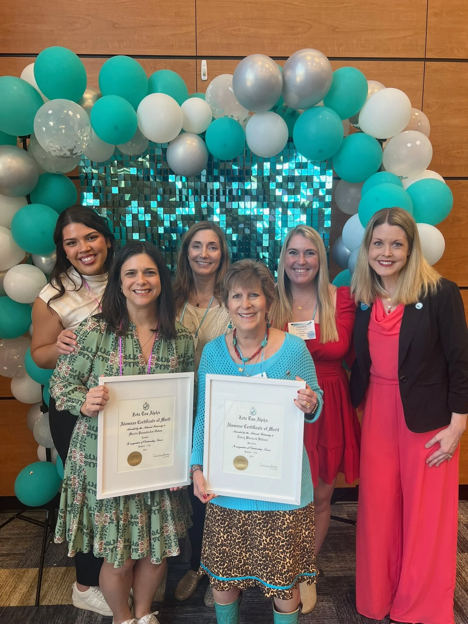 Six women standing in front of a balloon arch and a shiny, mosaic backdrop, with two women holding framed certificates of merit.