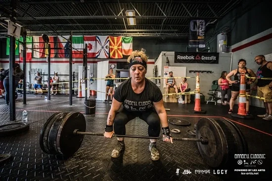 Woman in black athletic wear and headband lifting a heavy barbell in a CrossFit gym with flags hanging from the ceiling and other people working out in the background.