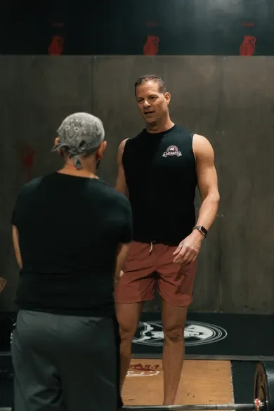 Two people having a conversation in a gym, one man wearing a black sleeveless shirt and red shorts, the other person with gray hair and a hat, with gym equipment and a concrete wall in the background.