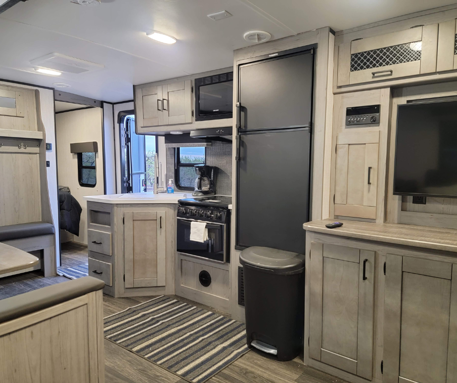 Interior of a compact RV with kitchen area featuring a microwave, refrigerator, stove, coffee maker, and TV, with light wood cabinets and a cozy seating area.