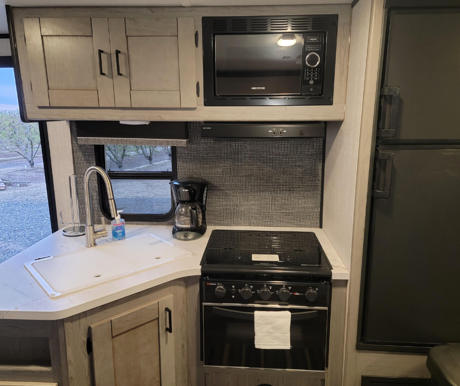 Compact kitchen corner with a white countertop, sink, coffee maker, microwave, stove, refrigerator, and window in an RV or small apartment.