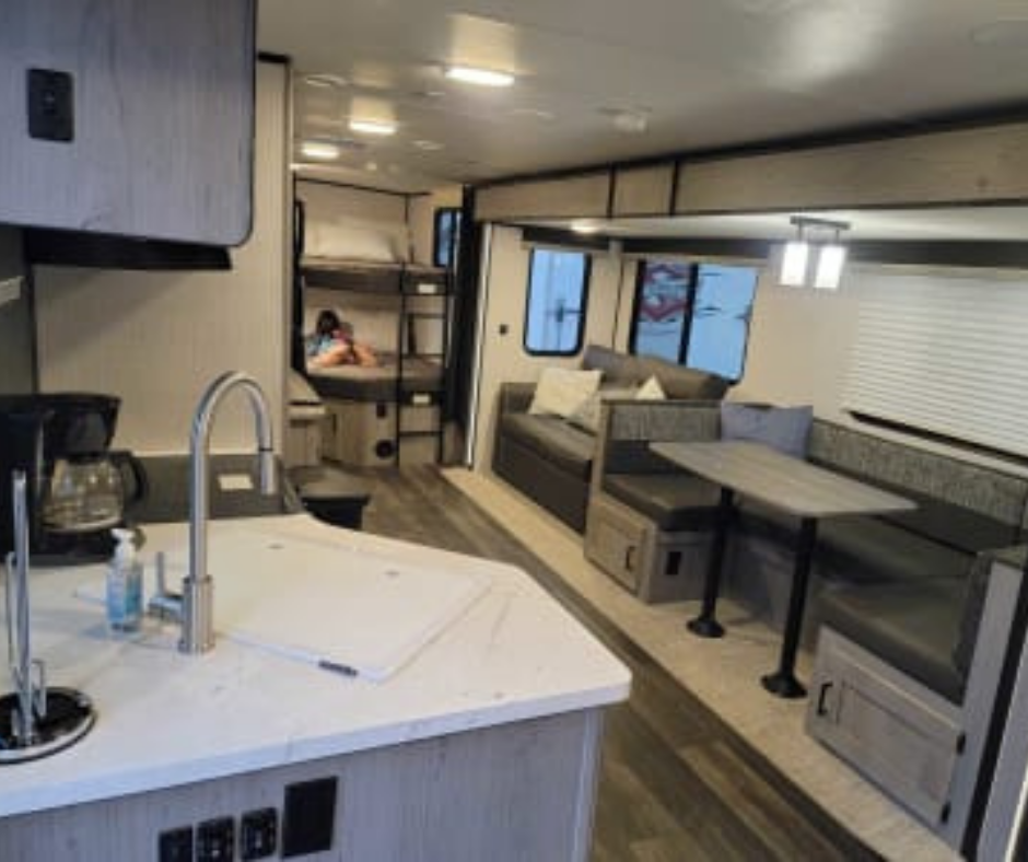 Interior of a modern RV with a kitchenette, a dining area with L-shaped seating, and a sleeping area with bunk beds, a person is sitting in the bunk bed area.
