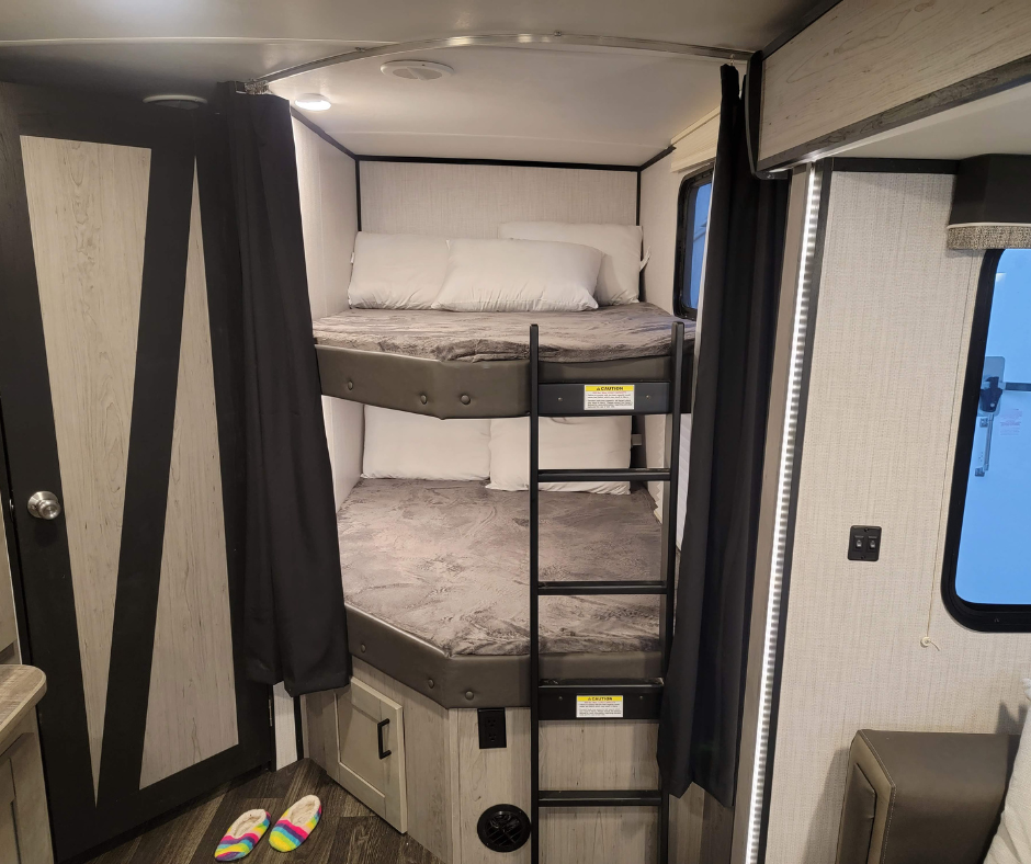 Bunk beds in a small RV or camper with black curtains and a window on the right side.