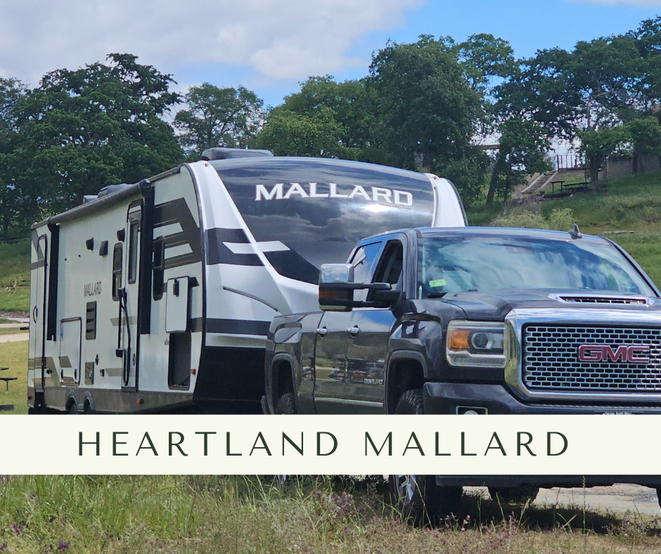 A black GMC truck attached to a white and black Heartland Mallard travel trailer parked on grass, with trees and a blue sky in the background.