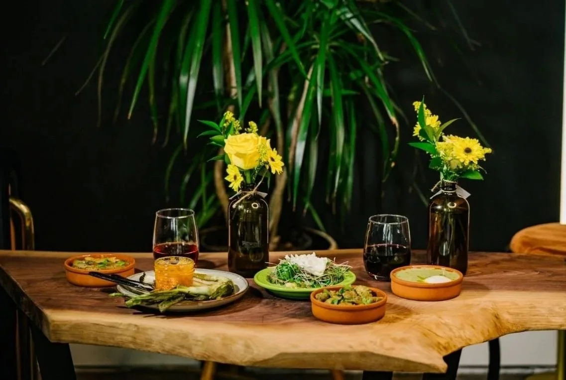 A wooden dining table set with four Spanish tapas in terracotta bowls of food, two glasses of red wine, and three small flower arrangements in dark vases, with a large green plant in the background.