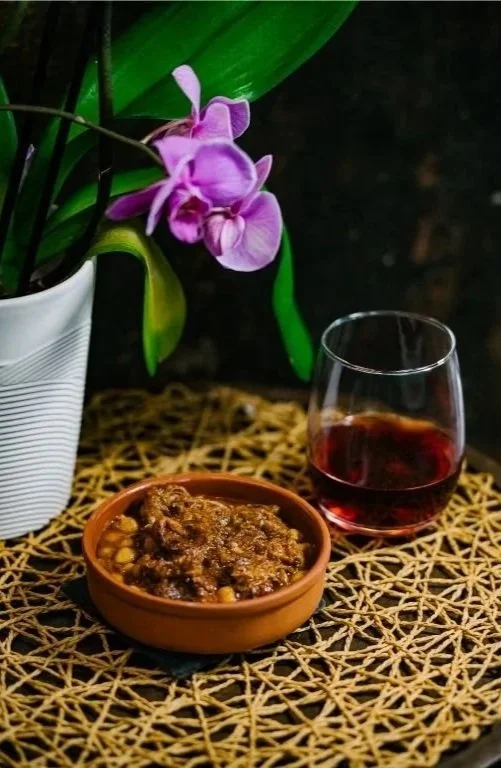 Purple orchid flower in a white pot, a glass of red wine, and a bowl of lamb and garbanzos (chickpeas) on woven placemat. Spanish tapas.