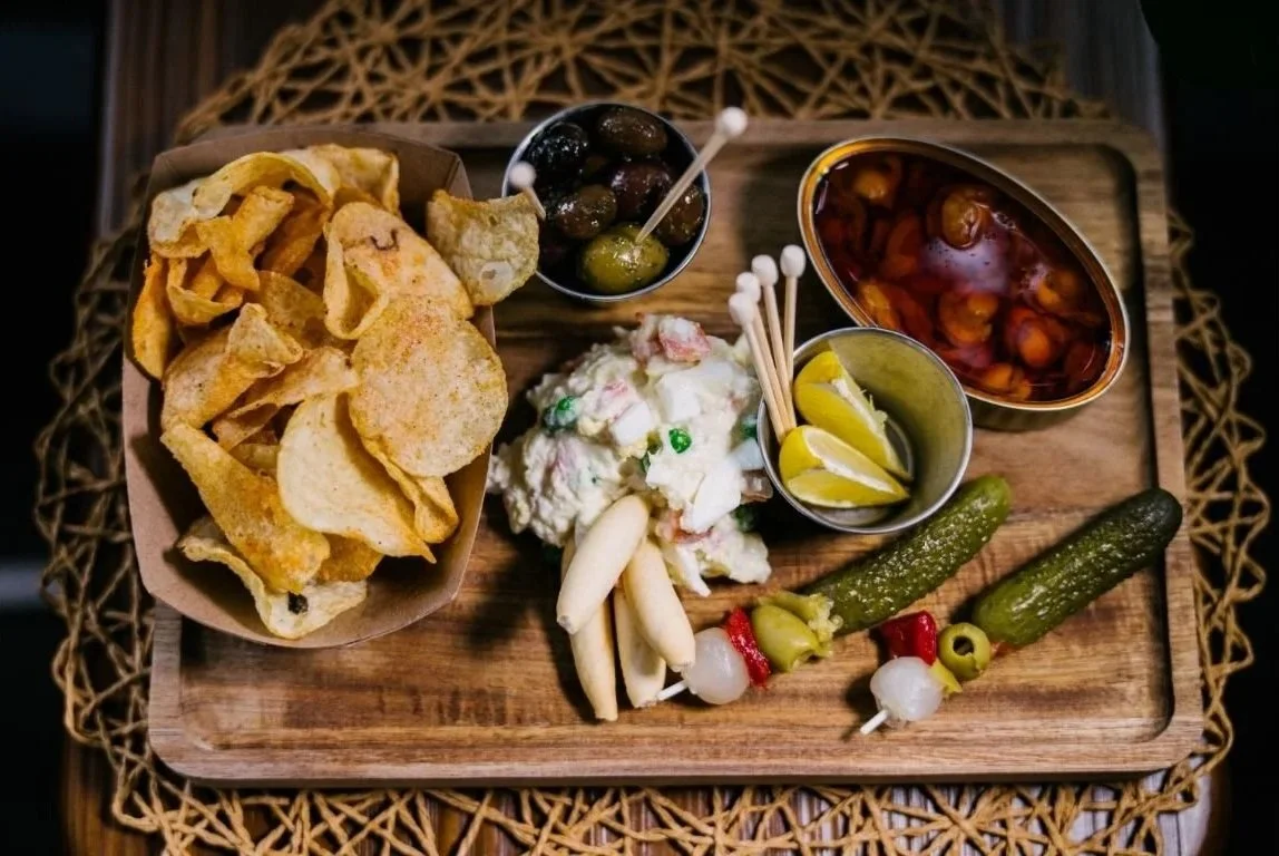 A wooden tray with potato chips, green and black olives, potato salad with peas and carrots, a bowl of pickled vegetables, and small skewers with pickles, onions, and peppers.