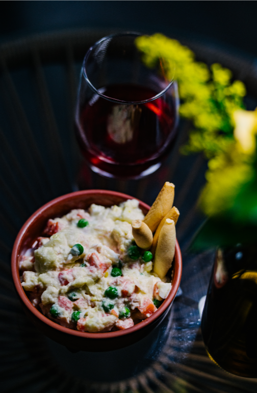 A bowl of potato salad topped with green peas and decorated with rolled wafer crackers, accompanied by a glass of red wine and a dark beverage, with yellow flowers in the background. Spanish tapa of ensaladilla rusa. 