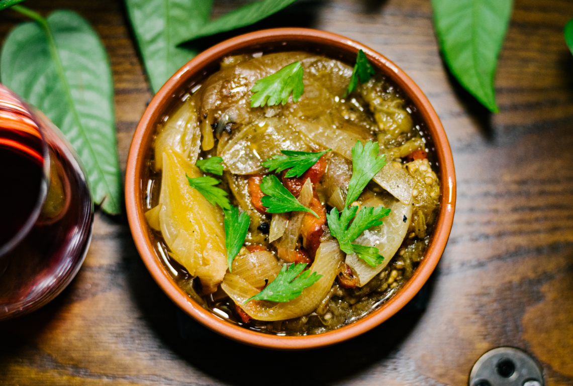 A bowl of cooked eggplant and vegetable stew garnished with parsley, with a glass of red wine on a wooden table and green leaves in the background. Spanish tapa of escalivada.