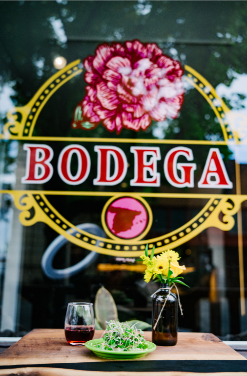 A bodega window with a painted logo of a pink flower, yellow circle, and the word 'Bodega' in red and white. On the table in front, there is a glass of red wine, a green plate of salad, and a dark bottle with yellow flowers. Spanish tapas.