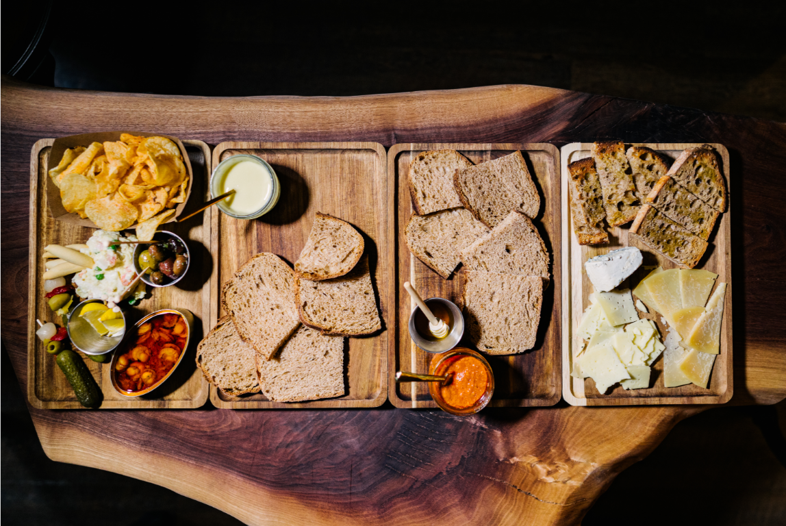 Wooden serving trays with potato chips, olives, pickles, red pepper spread, slices of bread, fresh bread, and cheese on a wooden table. Spanish tablas. Spanish tapas.