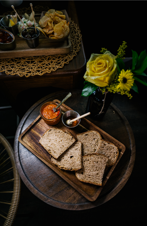 A wooden tray with slices of bread, a jar of honey, and a small bowl of sobrasada with a butter knife. A yellow flower arrangement is on a nearby table with snacks and drinks. Spanish tapas. Spanish tablas. Spanish board.