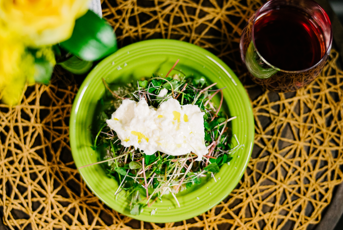 A meal with a white asparagus green salad topped with burrata cheese on a bright green plate, next to a glass of red wine on a woven wooden placemat. Spanish tapas.