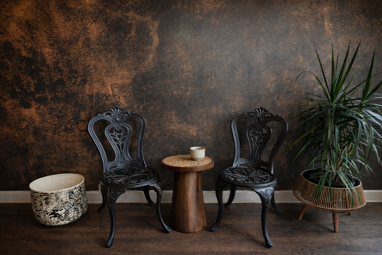 Two black vintage-style chairs with intricate designs, a small wooden table with a woven placemat and a cup, a large potted plant, and a decorative ceramic pot on a wooden floor against a textured dark brown wall.