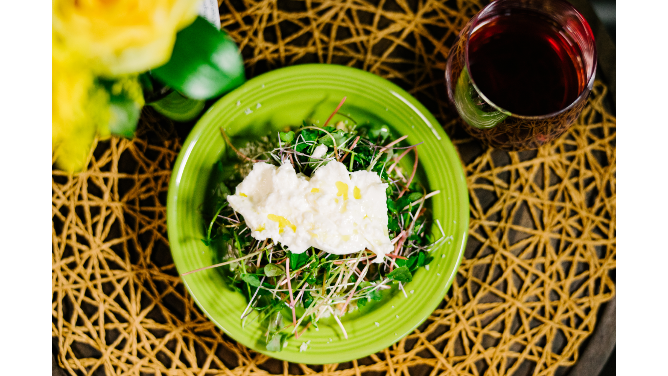 A meal with a white asparagus green salad topped with burrata cheese on a bright green plate, next to a glass of red wine on a woven wooden placemat. Spanish tapas.