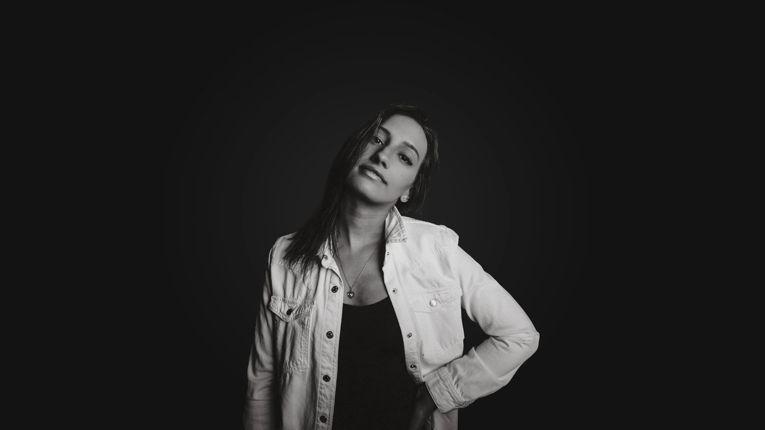 Black and white portrait of a woman with long dark hair, wearing a light-colored denim jacket over a black top, standing against a dark background with her left hand on her hip.