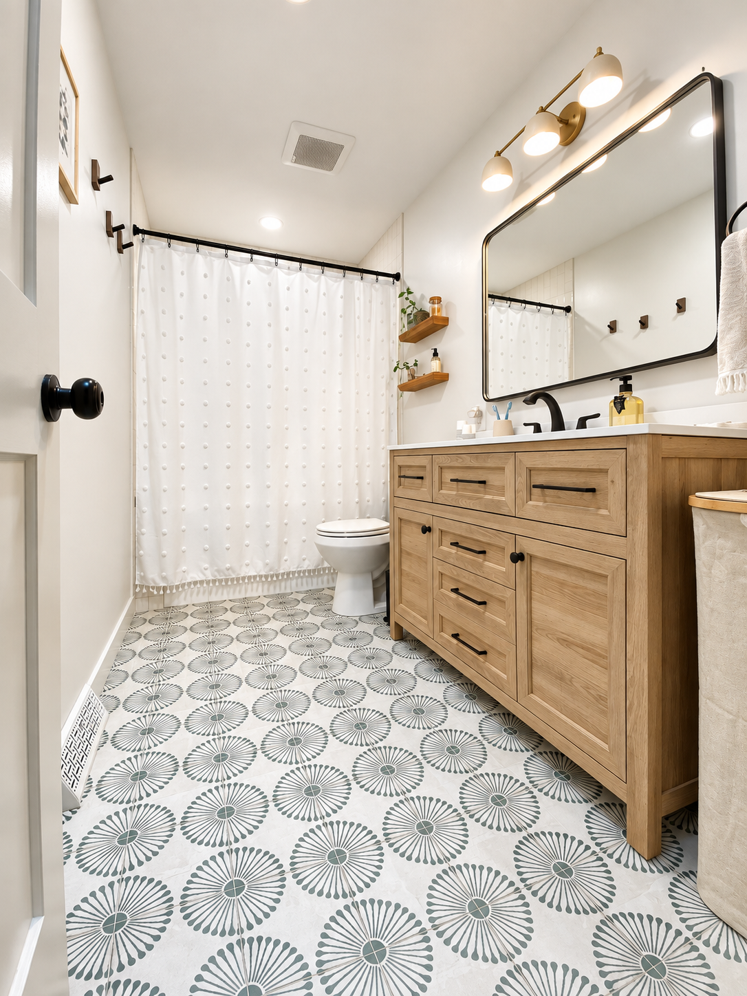 Modern bathroom with patterned tile floor, wooden vanity with black hardware, large mirror, white shower curtain, and small wall-mounted shelves with decorative plants and toiletries.