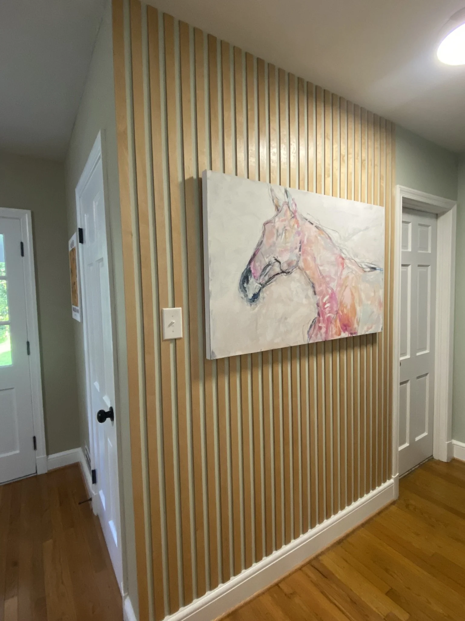 View of a wall with vertical wooden slats, a large abstract horse painting, a closed door on the right, and a partially visible door on the left, with hardwood flooring.