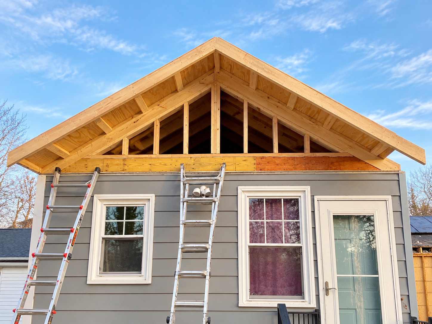 A house under construction showing the upper part with a roof frame, ladders leaning against the house, and windows on the lower part of the house, with a blue sky in the background.