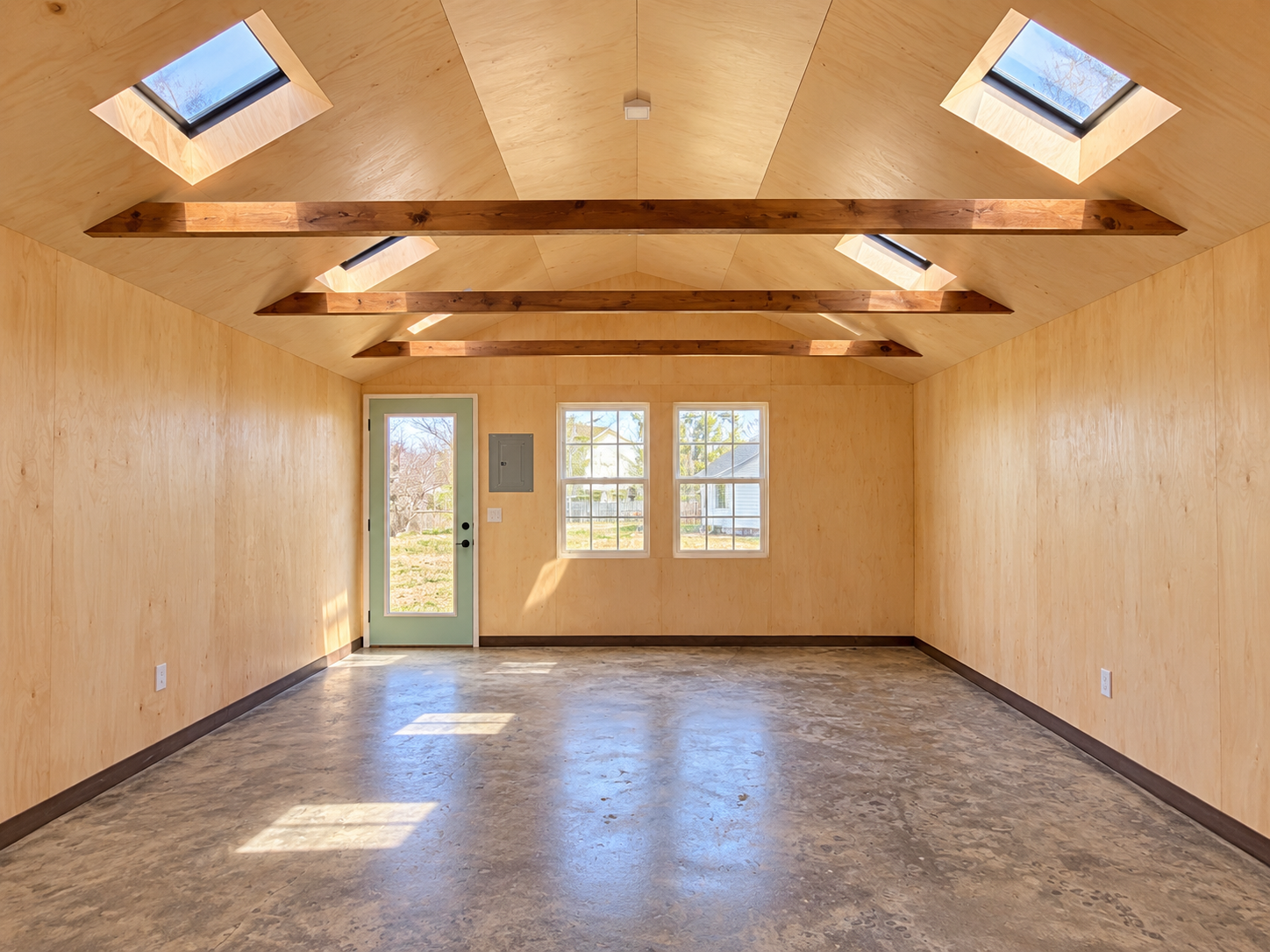 Empty room with wooden walls, ceiling, and beams, three skylights, two windows, a door, and a concrete floor.