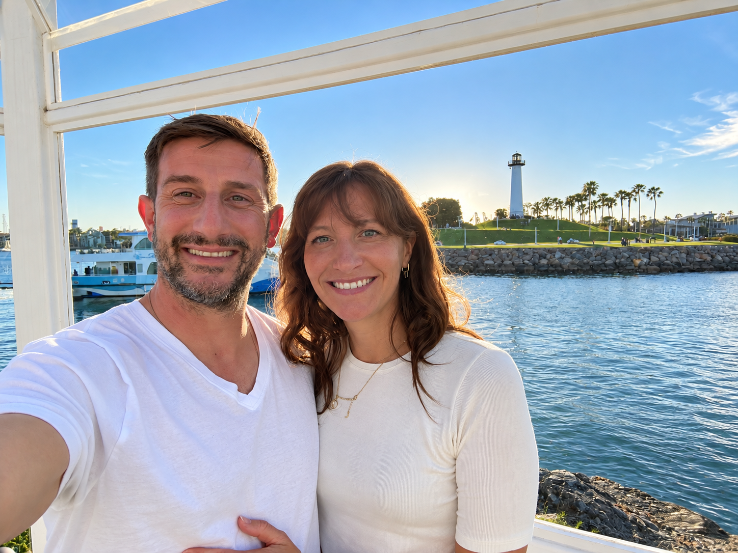 A smiling couple taking a selfie outdoors near a body of water with a lighthouse and palm trees in the background during sunset.