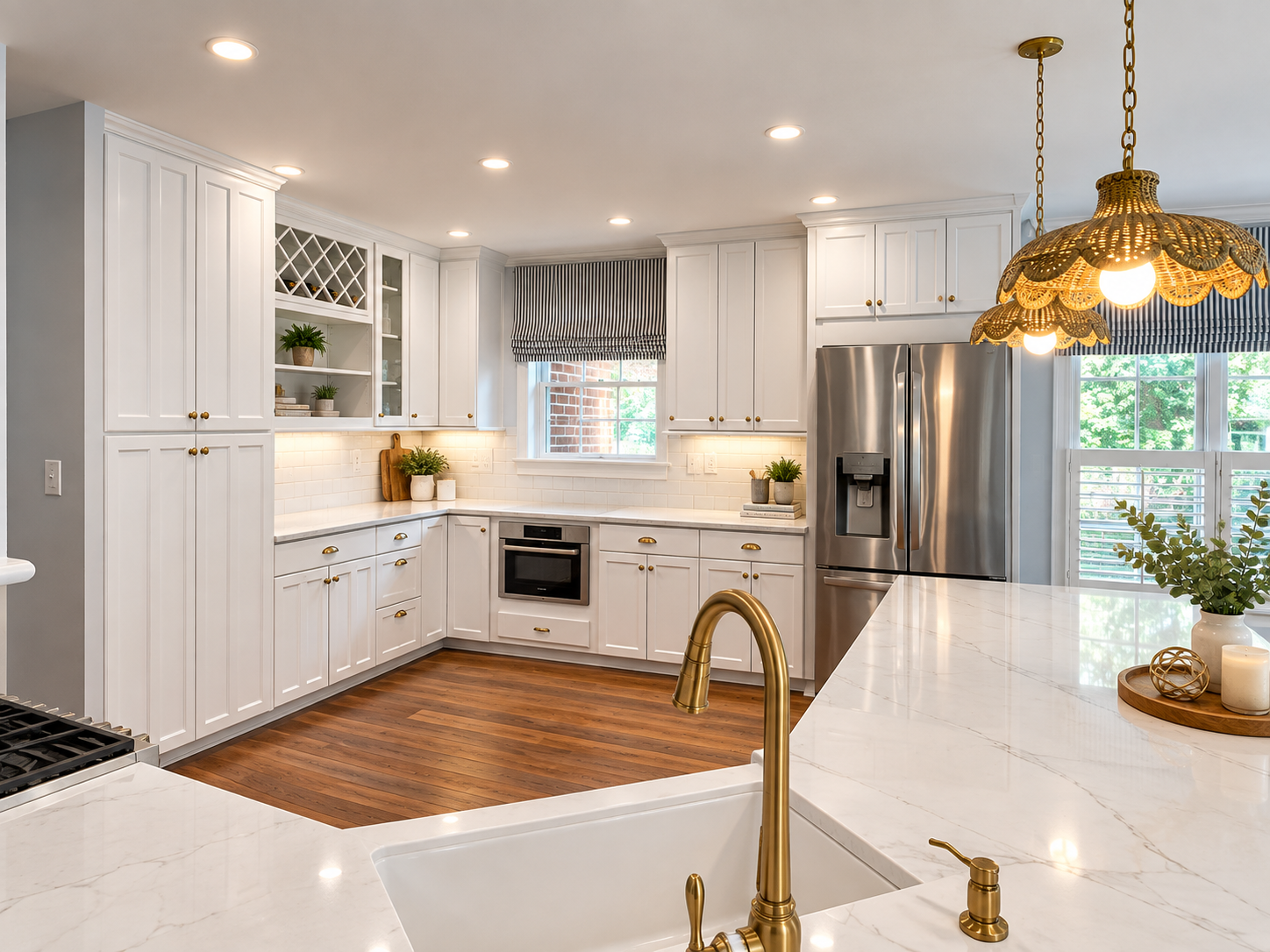 Modern kitchen with white cabinets, gold hardware, stainless steel appliances, a marble countertop island, and wooden flooring. Decorative pendant lights and window blinds add to the stylish decor.