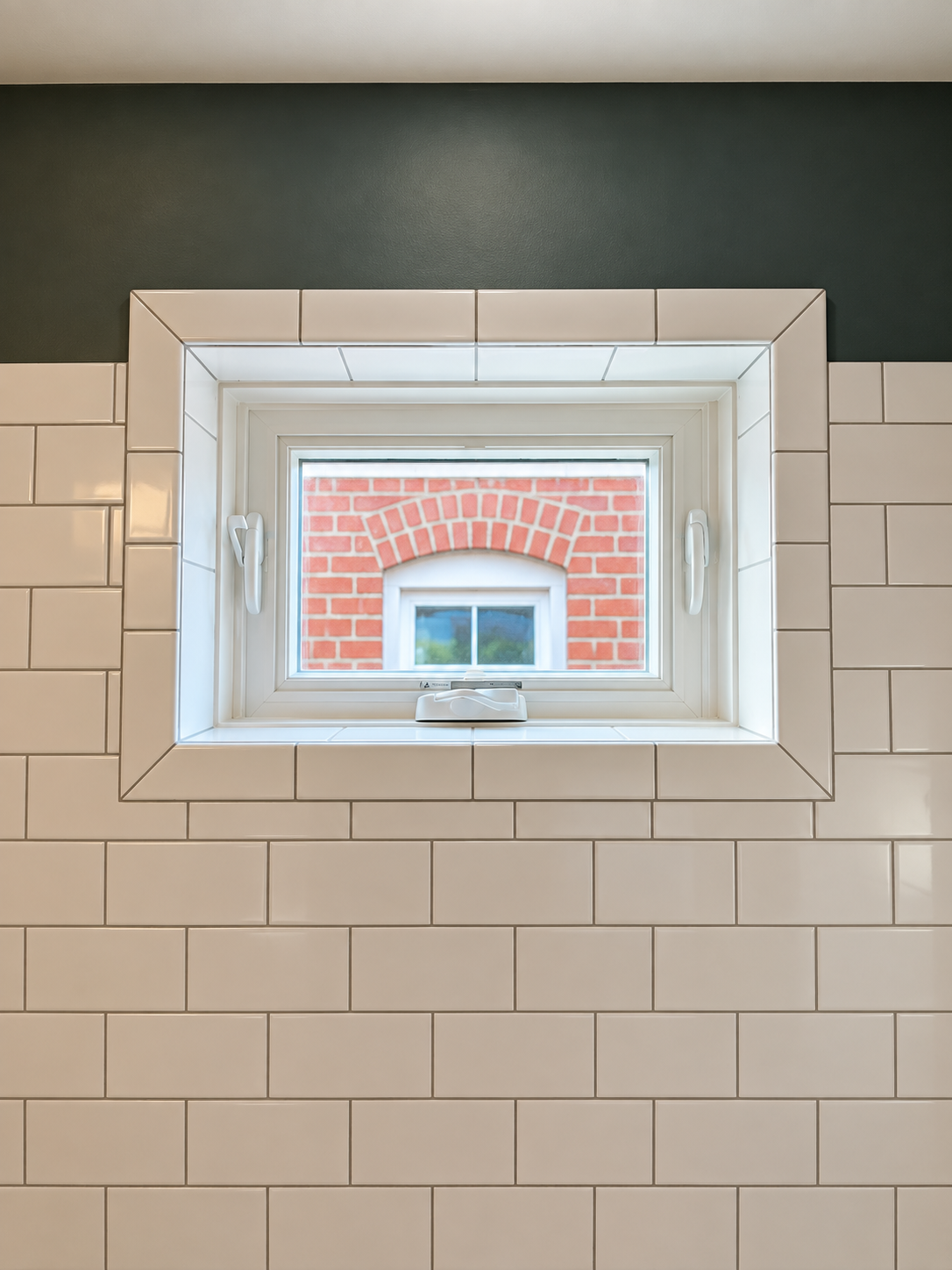 Close-up of a small rectangular window with white frame and latches, set in a wall decorated with white subway tiles, showing a view of a brick building with a small window and blue sky outside.