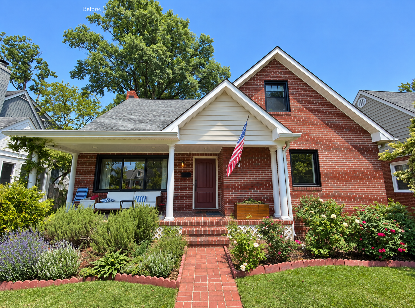 A brick house with a front porch, decorated with outdoor furniture, an American flag, and surrounded by a garden with colorful flowers and green bushes, under a clear blue sky.