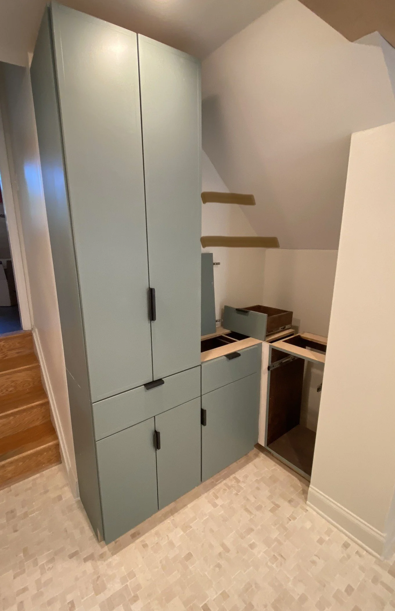 A partially assembled kitchen cabinet with multiple drawers and doors, with some parts still missing or unassembled, on a tiled floor in a room with beige walls.