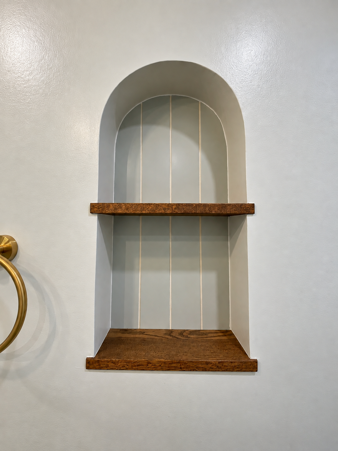 Empty built-in wall shelf with three wooden tiers, arched top, and white vertical panel backing, mounted on a white wall with a gold towel ring visible on the left side.