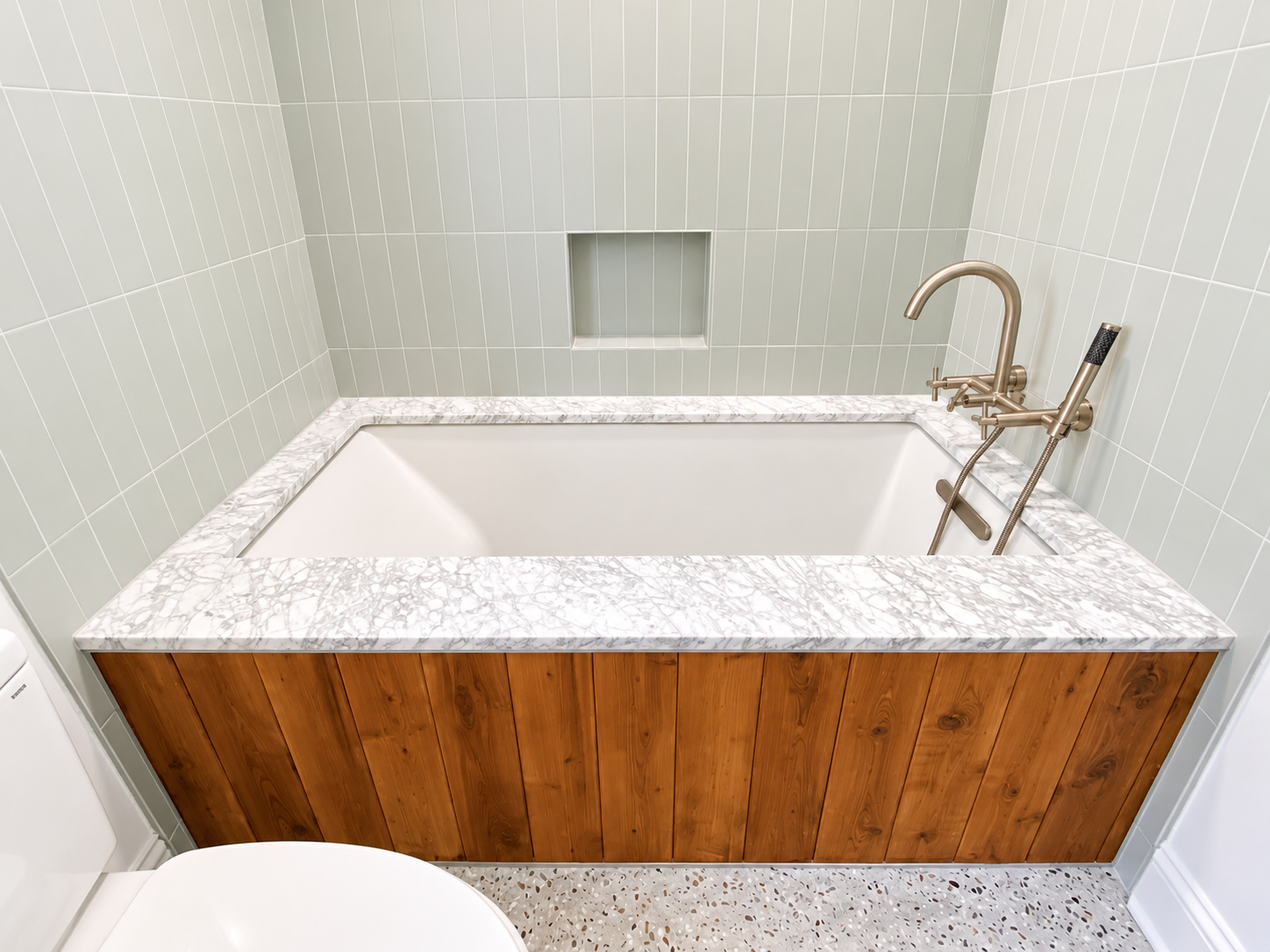 A modern bathtub with a marble surround and wooden panel exterior, set against light green tiled walls. There is a built-in niche in the wall above the bathtub, and a gold-tone faucet with a handheld shower attachment.