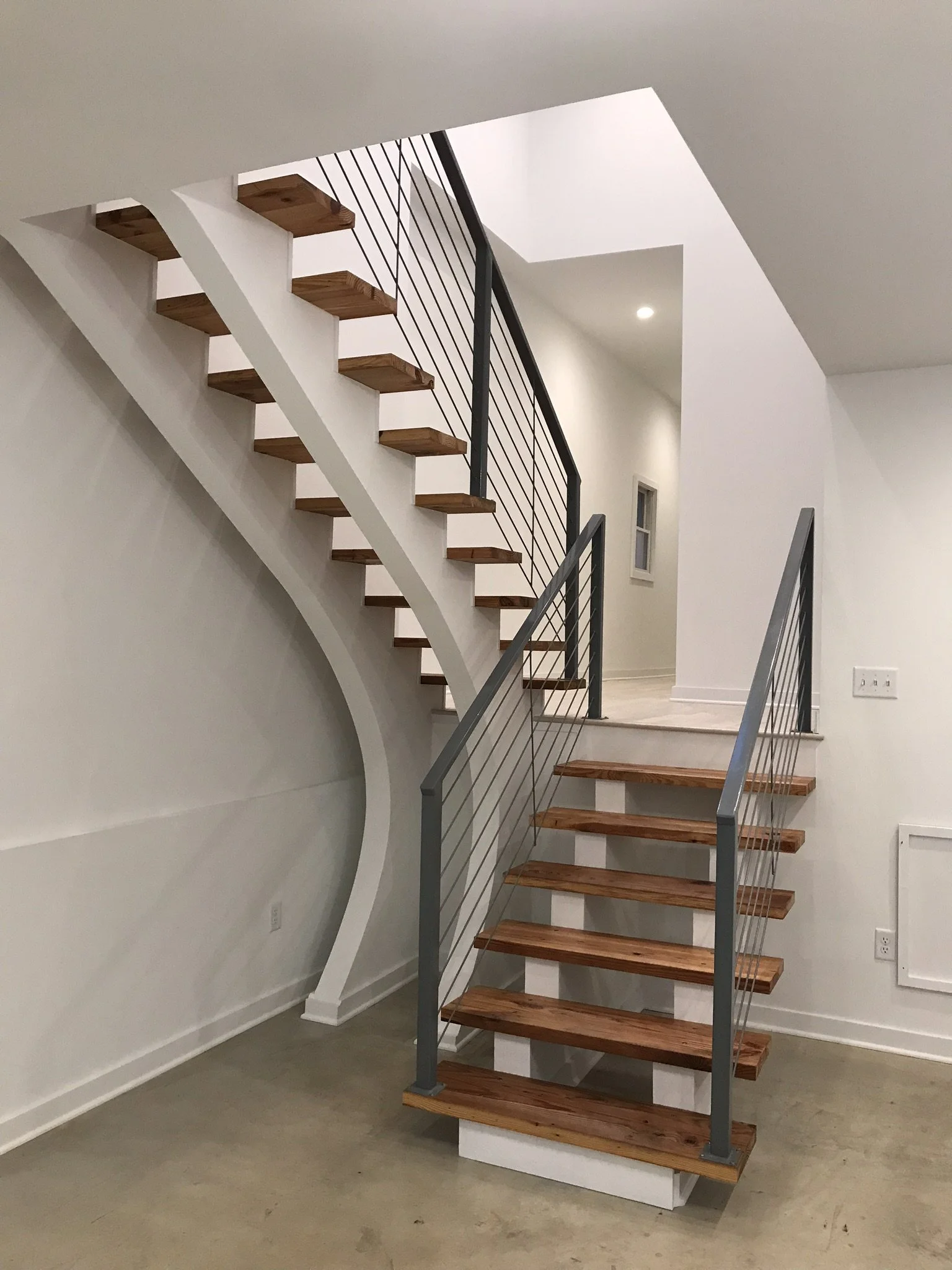 Interior view of a modern staircase with wooden steps, white sides, and metal railings, leading to an upper floor in a contemporary home.