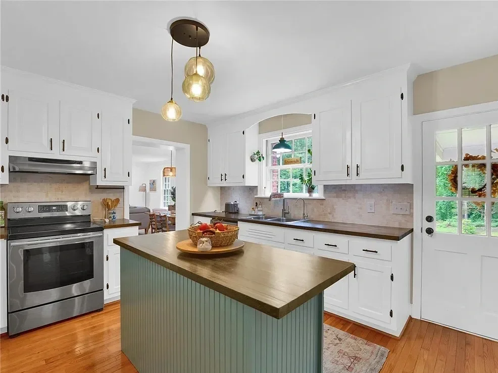 Bright kitchen with white cabinets, a wooden countertop, and a small central island with a green base. Stainless steel oven, a window above the sink, and a door with glass panes on the right.