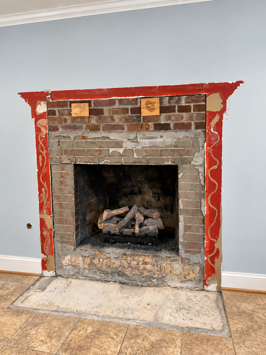 In-progress fireplace renovation with exposed brick and drywall, firewood inside, torn wallpaper or wall covering, on a tiled floor.